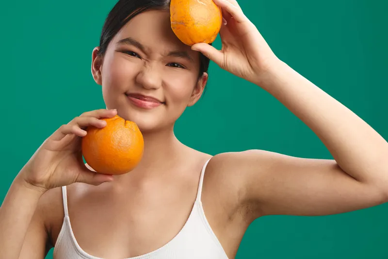 cheerful asian woman holds oranges in a bright studio, smiling toward the camera, creating a fresh healthy lifestyle mood with vibrant colors and a friendly, relaxed vibe.