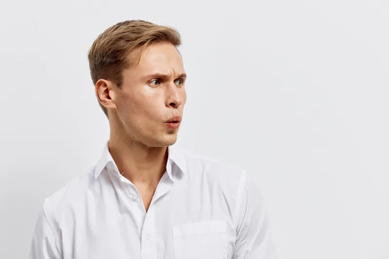 Thoughtful young man with short brown hair wearing a white shirt, looking to the side with a curious expression, isolated on plain white background, studio portrait. People lifestyle concept