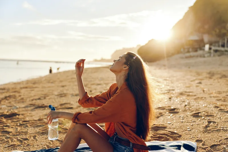 Woman beach sunset water bottle sitting blanket ocean nature relaxation, relaxing outdoors, holding water bottle, drinking on sand shore, warm golden sunlight, vacation mood by the sea