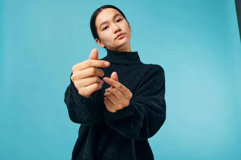 Young woman in black sweater posing with hand gesture on blue background. Female model looking confident and stylish with casual outfit in studio lighting.