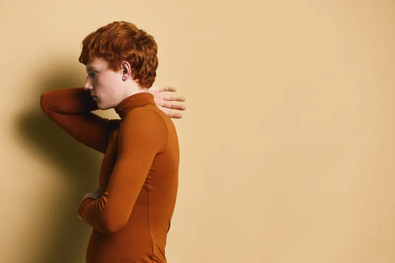 Redhead woman in brown turtleneck standing sideways with arms crossed on beige background. Female model with short hair and thoughtful expression in studio portrait with simple backdrop.