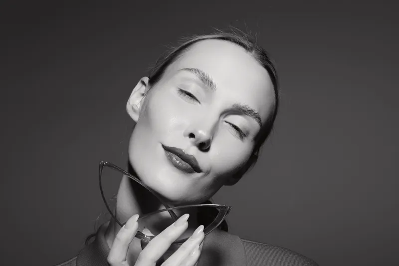 Close-up portrait of a young white woman with light skin holding sunglasses near her face with closed eyes, expressing calm and elegance in studio lighting.