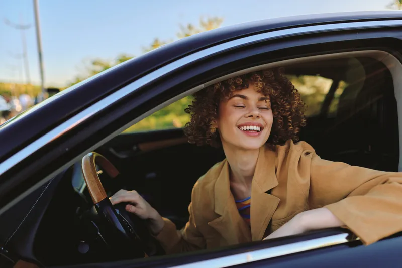 Woman driving a car, smiling with curly hair, wearing a casual coat, captured in warm retro film color, lifestyle portrait with sunlight, cheerful road moment, candid and relaxed