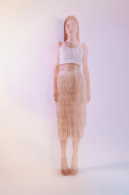 Young woman in a white crop top and beige fringe skirt stands against a soft pastel background. Concept for beauty, fashion, and elegance in modern style.