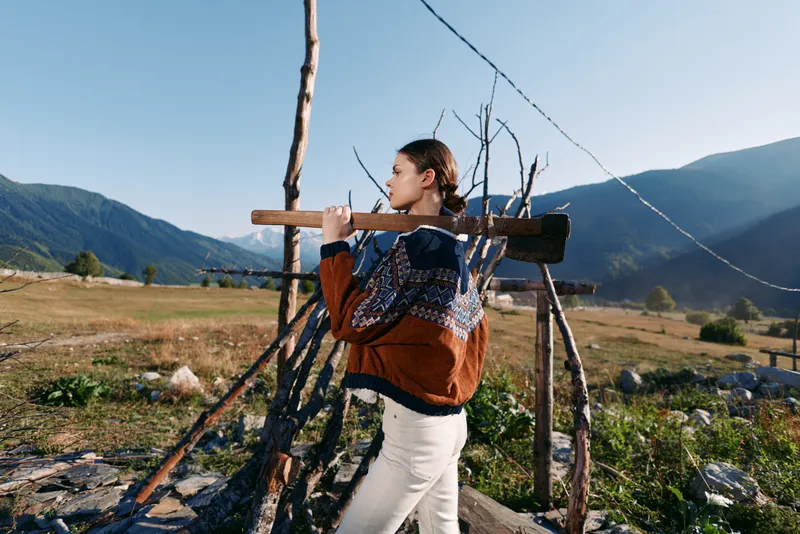 Woman carrying wood on rustic fence in mountains, wearing a warm sweater in a rural landscape; portrait of outdoor activity and traditional lifestyle on a sunny day.