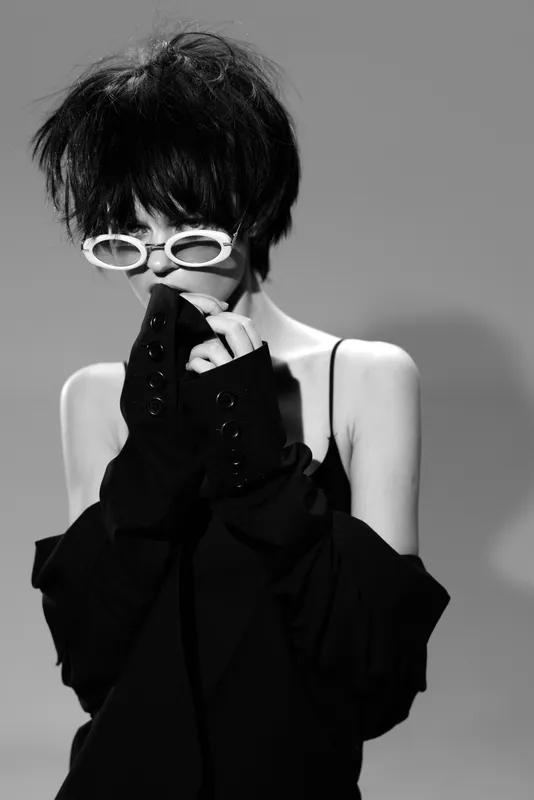 Black and white portrait of a woman with short hair wearing white glasses and dark clothing, expressing mystery and elegance.