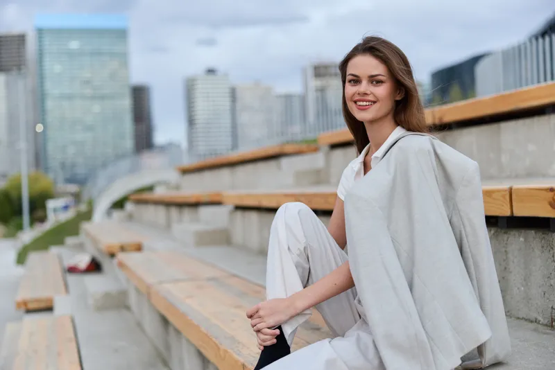 Smiling woman in light casual outfit sitting on wooden benches with urban cityscape background, outdoor lifestyle, modern fashion concept