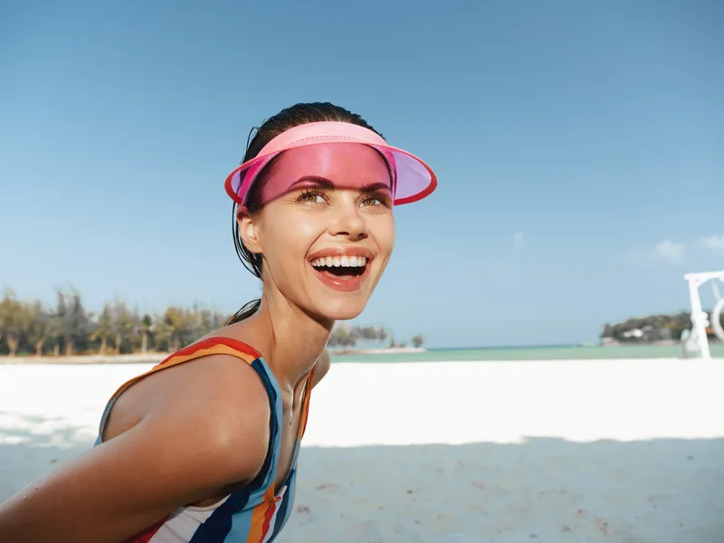 Joyful woman on a sunny beach, wearing a colorful swimsuit and pink visor, smiles toward the camera as white sand and turquoise water frame her energetic pose.