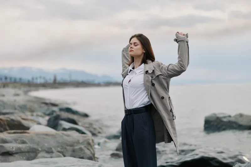 Young woman with closed eyes wearing grey coat and white shirt stands relaxed on rocky beach with sea and cloudy sky in background. Concept of freedom, calm, and nature connection.