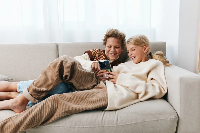 Authentic lifestyle scene shows a couple relaxing on a sofa, sharing a playful moment while using a phone together, in soft natural light and cozy home atmosphere.
