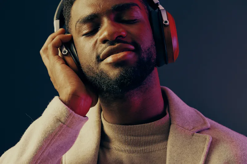 Young Black man enjoying music with closed eyes, wearing stylish headphones and a beige jacket against a dark blue background, conveying relaxation and joy