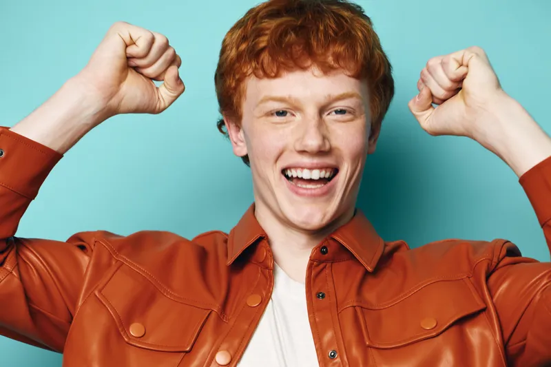 Happy man with red hair and freckles wearing brown jacket smiling and raising fists in celebration against blue background in studio portrait.