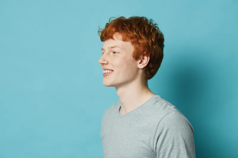 Young man with red hair and freckles smiling in profile portrait against blue background wearing casual grey shirt with positive expression and relaxed posture.