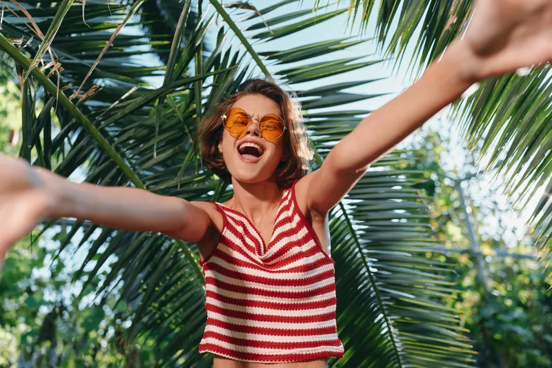 A cheerful young woman wearing a red striped top and sunglasses joyfully outdoors, arms lifted, sunshine and palm trees around, expressing freedom, happiness, and carefree summer energy.