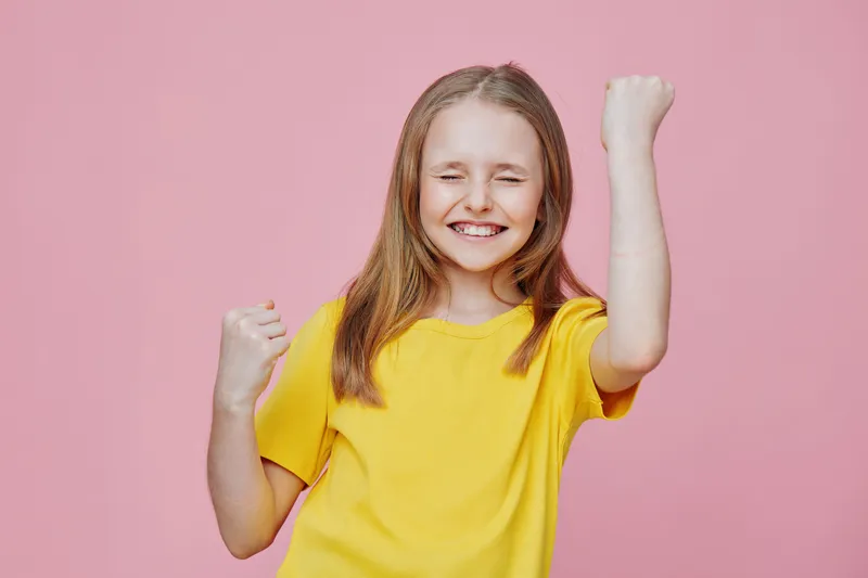 Happy young girl in a yellow t shirt celebrating against a pink background, expressing joy and positivity with a victorious gesture and bright smile