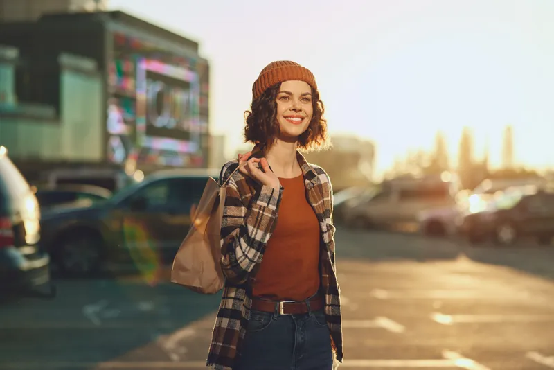 woman smiling golden candid urban lifestyle beanie bag. Young female portrait in parking lot at golden hour glow, authenticity and mindful living captured in warm sunlight and emotional storytelling.