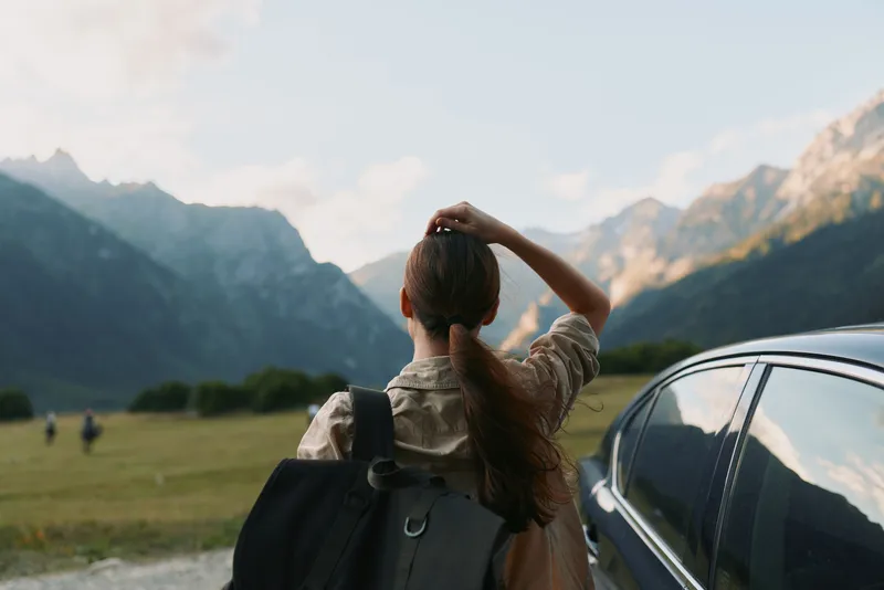 Woman with backpack standing beside a parked car, gazing at distant mountains, enjoying outdoors lifestyle, ready for travel and adventure in a scenic valley landscape.