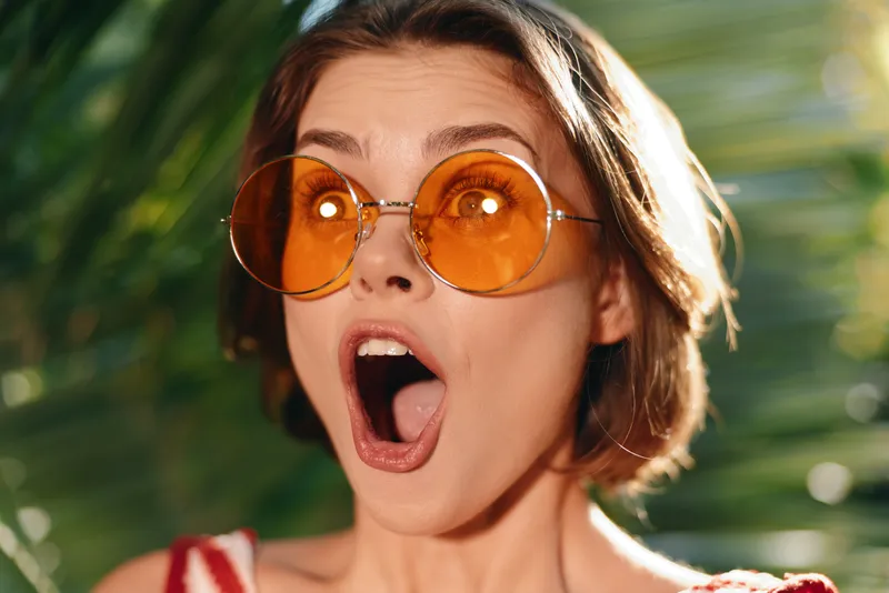A surprised woman with round orange sunglasses looks upward in a sunny outdoor setting, conveying astonishment and curiosity while creating a playful, vibrant mood.