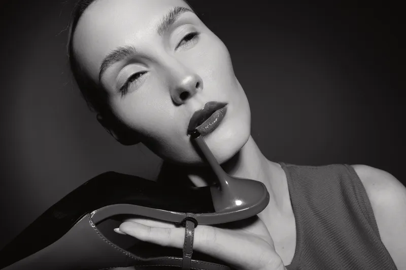 Close-up of a pale white woman holding a black high heel shoe near her lips with closed eyes in studio lighting.
