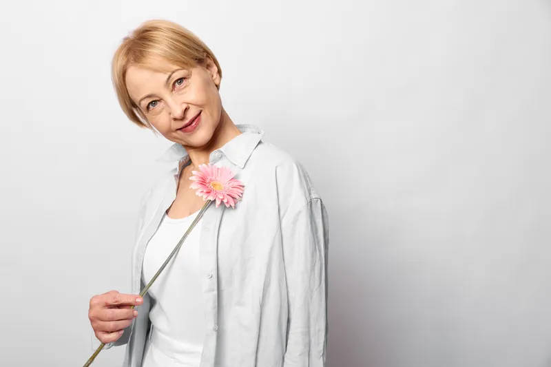 A woman doctor in white coat, stethoscope around neck, holds a flower, pink bloom, healthcare concept for physician compassion, calm smile and wink, bright studio background for wellness