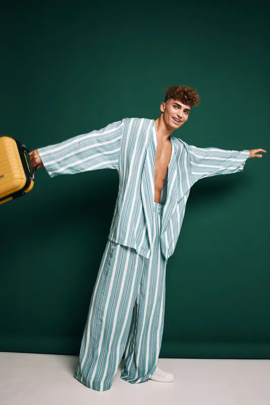 Man wearing colorful striped oversize pajamas smiling and holding a travel suitcase with open arms against green background, fashion style, casual comfortable clothing.