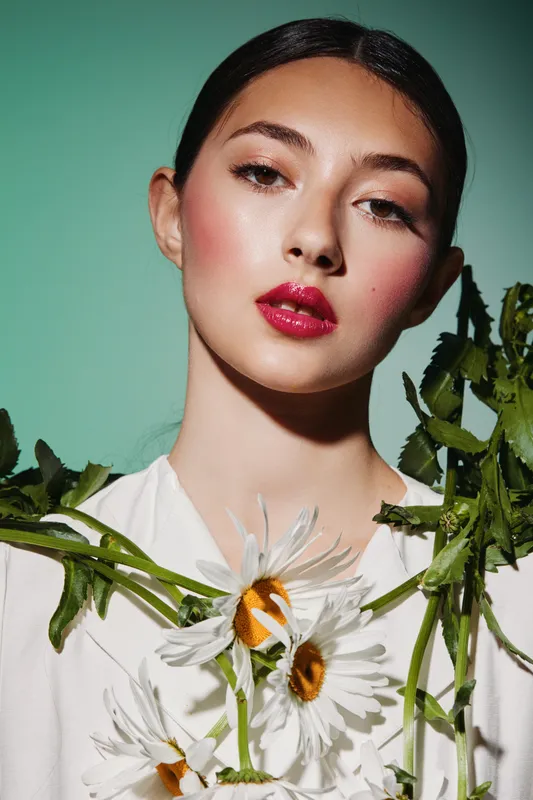 Young woman with glossy pink lips and dewy skin poses with fresh chamomile flowers on her shoulders against a light green background. Concept for beauty and fashion creativity.