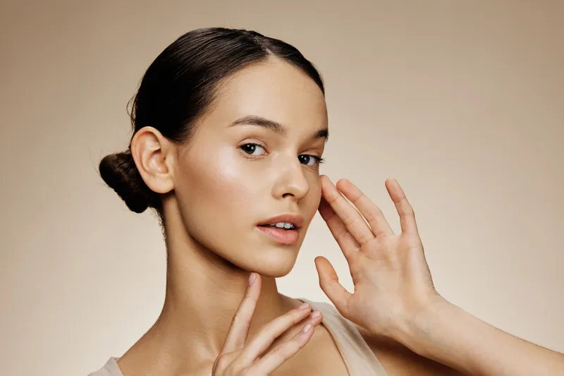 Young model with flawless skin showcasing natural beauty, wearing minimal makeup and gentle smile, against a warm beige background, emphasizing skincare and self care