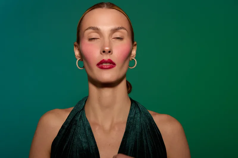 Portrait of a woman with closed eyes wearing dark green dress and gold hoop earrings on green background. Concept of elegance, fashion, beauty, and calm expression in studio lighting.