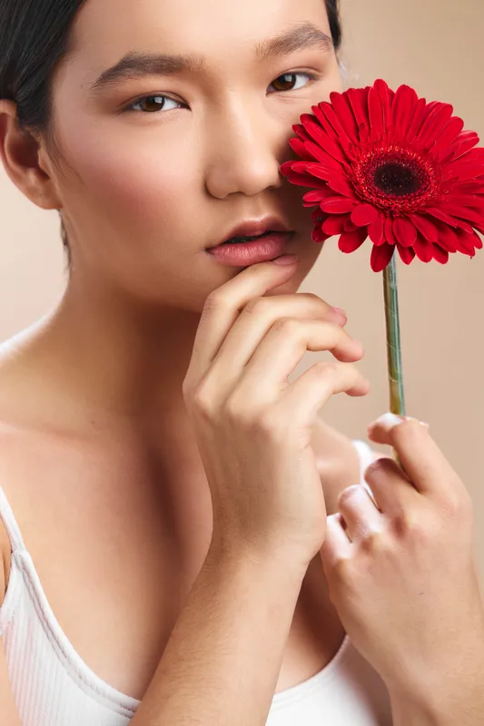 Asian woman gently holds a bright red flower near her face, showcasing calm elegance and soft lighting in a minimal studio setting.