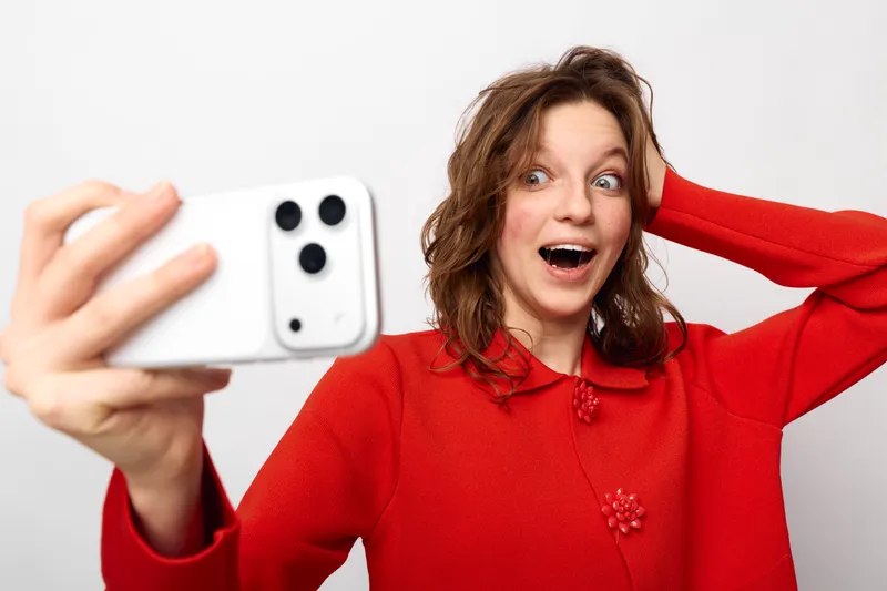 Surprised young woman in red sweater takes selfie with smartphone on white background. Concept of social media, communication, technology, and casual lifestyle with emotional expression.