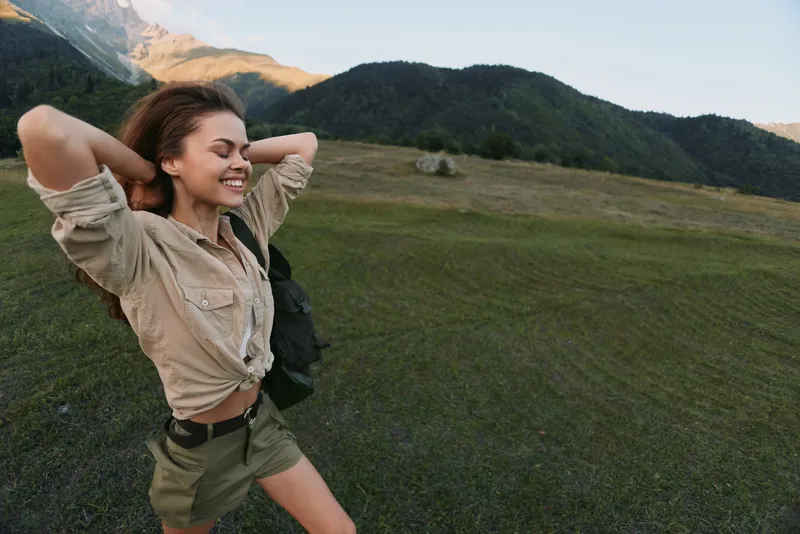 A young woman outdoors in a grassy valley stretches with hands behind her head, enjoying the fresh air and distant mountains.