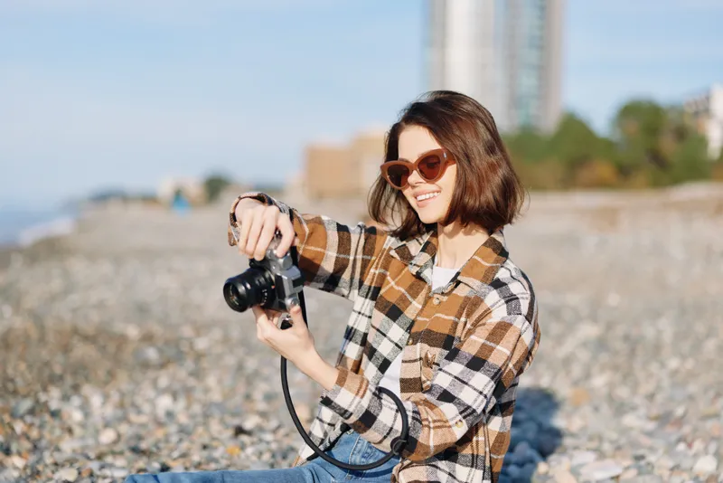 Photographer on the beach with a camera, wearing plaid shirt and sunglasses, capturing candid moments under bright sun for a relaxed outdoor lifestyle image