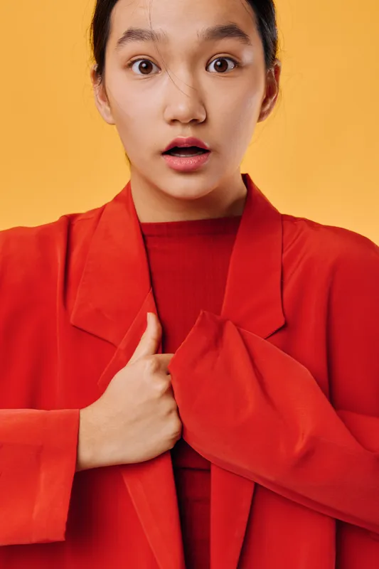 Young woman with shocked facial expression wearing red jacket and shirt, holding her chest, standing against orange background in casual studio portrait with copy space.