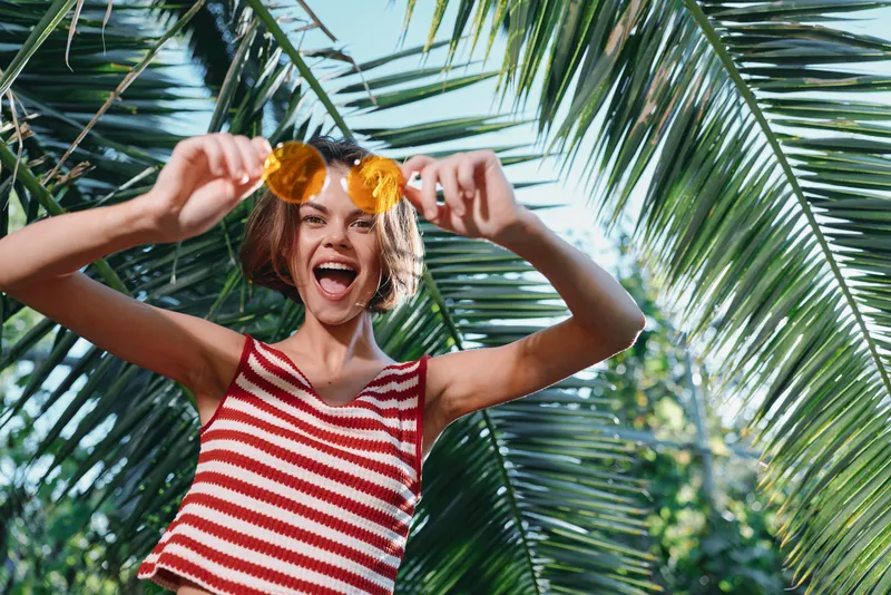 A cheerful woman stands outdoors among palm fronds, wearing a red and white striped top and sunglasses, smiling broadly as she enjoys a sunny, carefree moment.