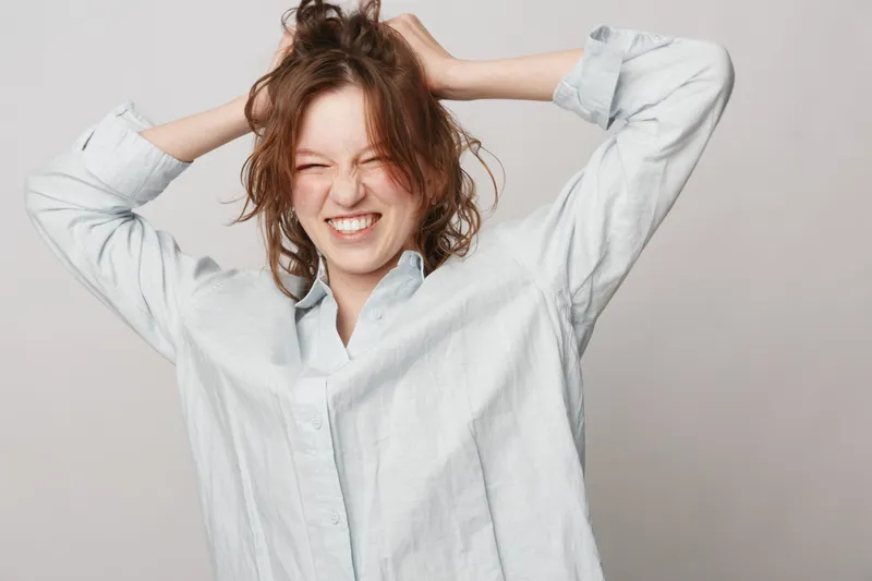 Young woman in casual shirt pulls her hair with eyes closed and teeth clenched. Emotional expression of frustration or stress on neutral background. Concept of tension and anxiety.
