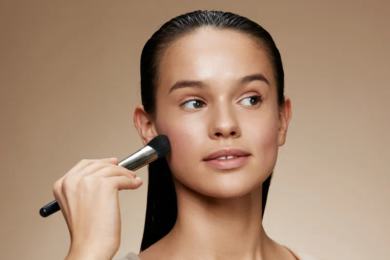 Young woman applying makeup with a brush, showcasing a natural look and glowing skin, perfect for beauty and skincare themes