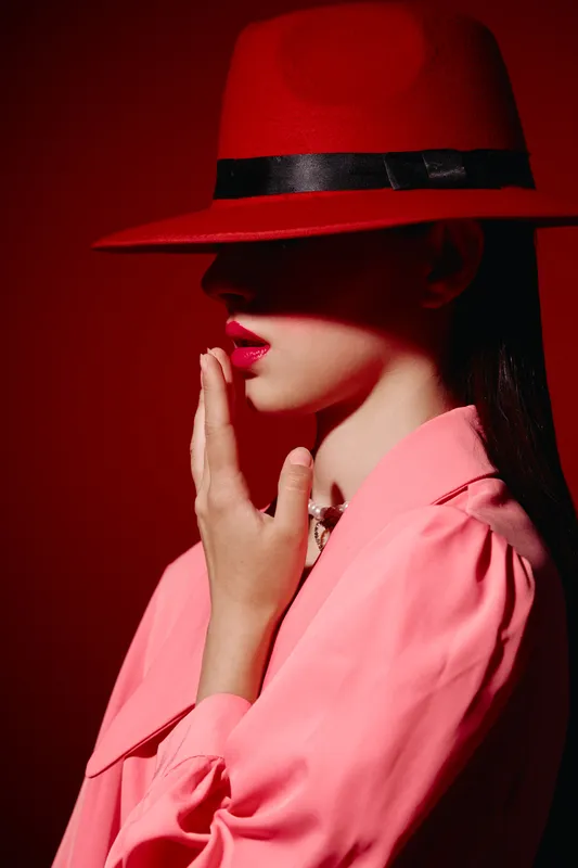 Portrait of woman in dramatic red hat and pink blouse with bold lipstick. Shadowed face creates mystery. Concept for fashion, beauty, mood, and editorial creative.