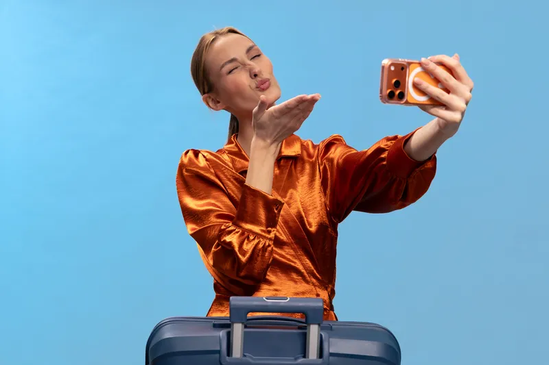 Young woman in shiny orange blouse takes selfie while blowing a kiss. She stands behind blue suitcase on blue background. Concept of travel, vacation, social media, and communication.