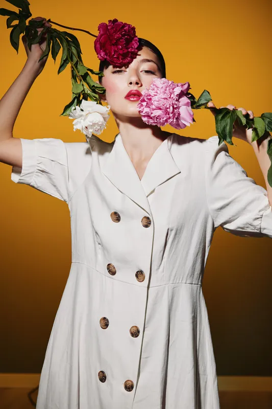 Young fashionable woman with red lipstick wears white buttoned dress and holds branch with pink and white flowers near her face on warm yellow background. Beauty and creative fashion concept.