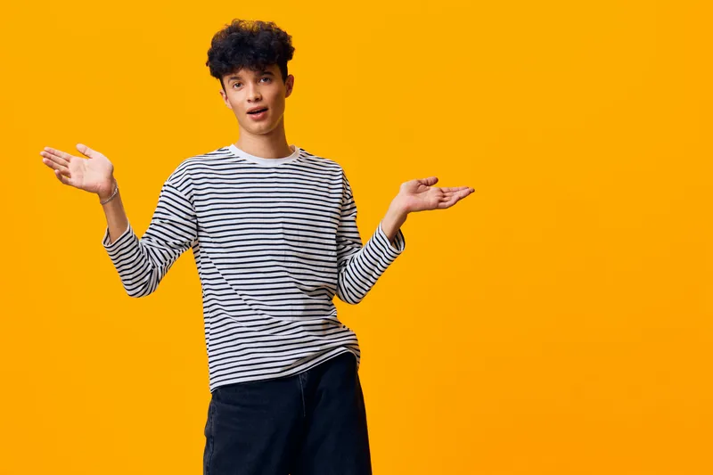 young man in striped shirt with curly hair shrugging hands against bright orange background expressing uncertainty or confusion in casual wear