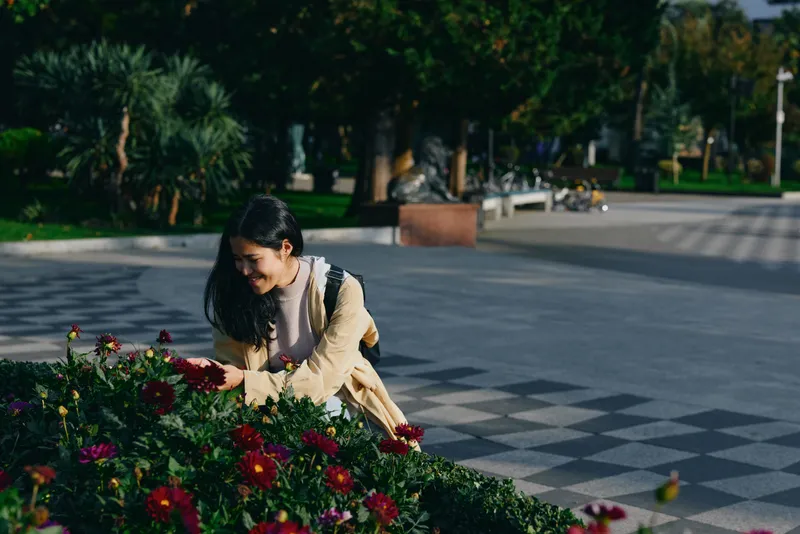 A woman bends near a bed of colorful flowers in a sunlit park, admiring blooms, tending a garden path with a calm, focused expression, enjoying outdoor quiet and gentle nature moments.