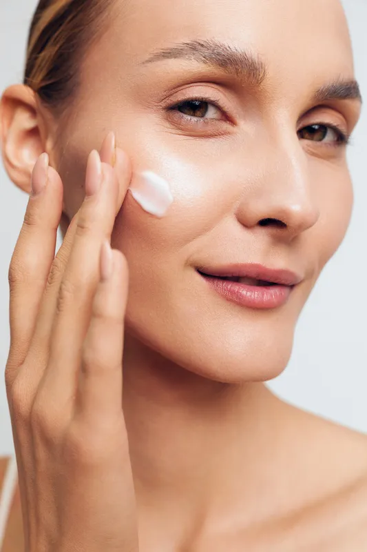 Young woman with clear skin applies moisturizing cream on her cheek in a close-up beauty portrait with natural light and a soft expression.