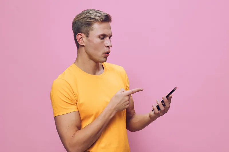 Young man wearing yellow t-shirt using smartphone pointing at screen isolated on pink background studio portrait. People lifestyle concept, technology, communication, digital device, casual style, scr