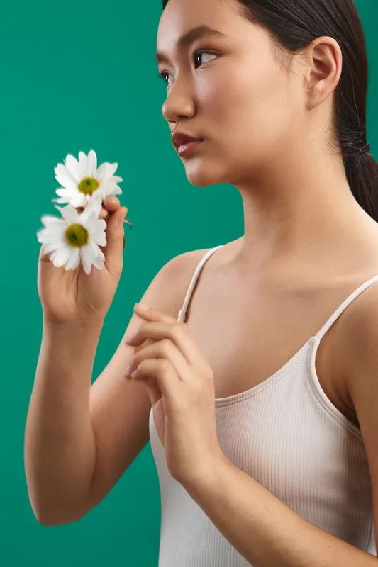 Asian woman holds a bouquet of daisies against a teal background, calm and graceful pose, soft lighting, conveying freshness and gentle focus.