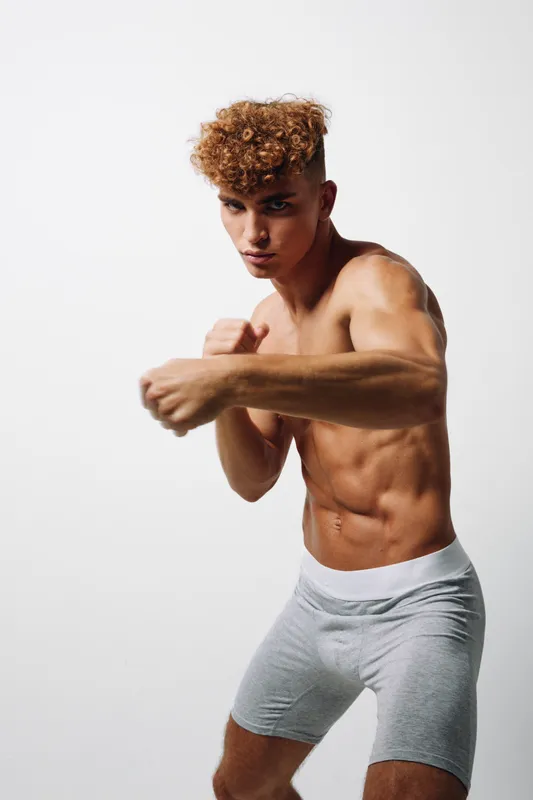 Athletic young man with curly hair practicing boxing punch in gray shorts against white background. Muscular fit male training for fitness and combat sport workout.
