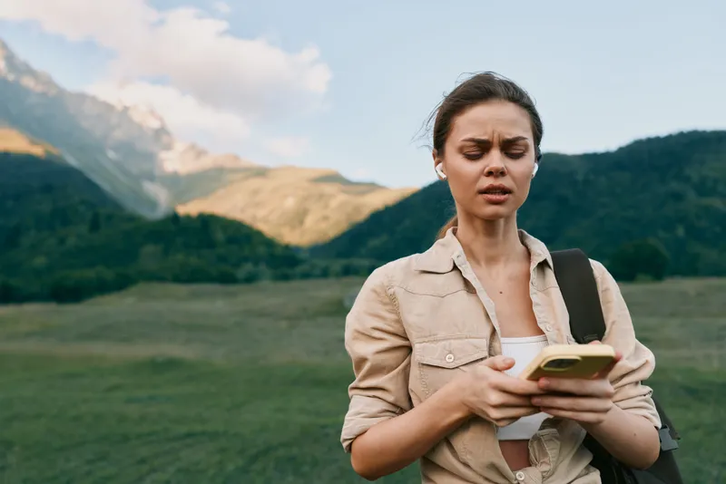 Young woman with brunette hair checks her smartphone outdoors in a mountainous area during daytime, showing a focused and thoughtful expression.
