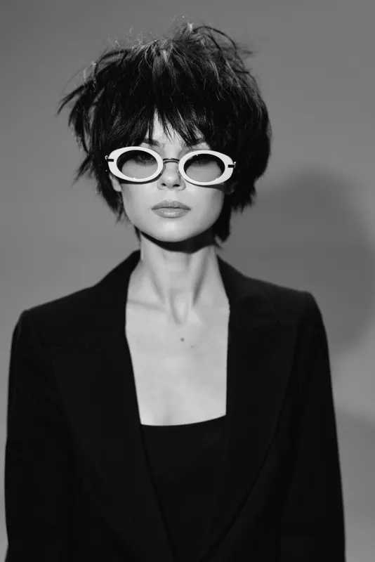 Stylish woman wearing white sunglasses and a black blazer in a black and white portrait with a modern hairstyle.