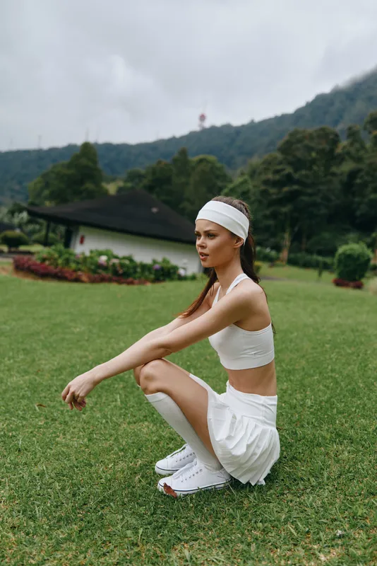 Young athletic woman in a white sports outfit squats outdoors surrounded by greenery, showcasing a fit lifestyle and determination