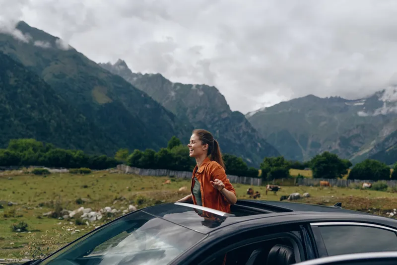A woman stands beside a sleek car in a dramatic mountain landscape, enjoying the breeze and wide open scenery during a scenic road trip