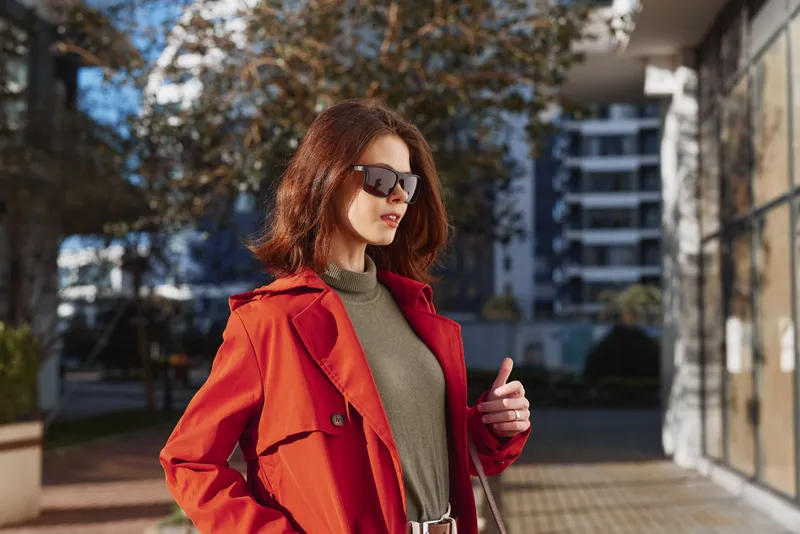 Stylish woman in a red coat and sunglasses stands on a city street, posing confidently with a hand on her belt.